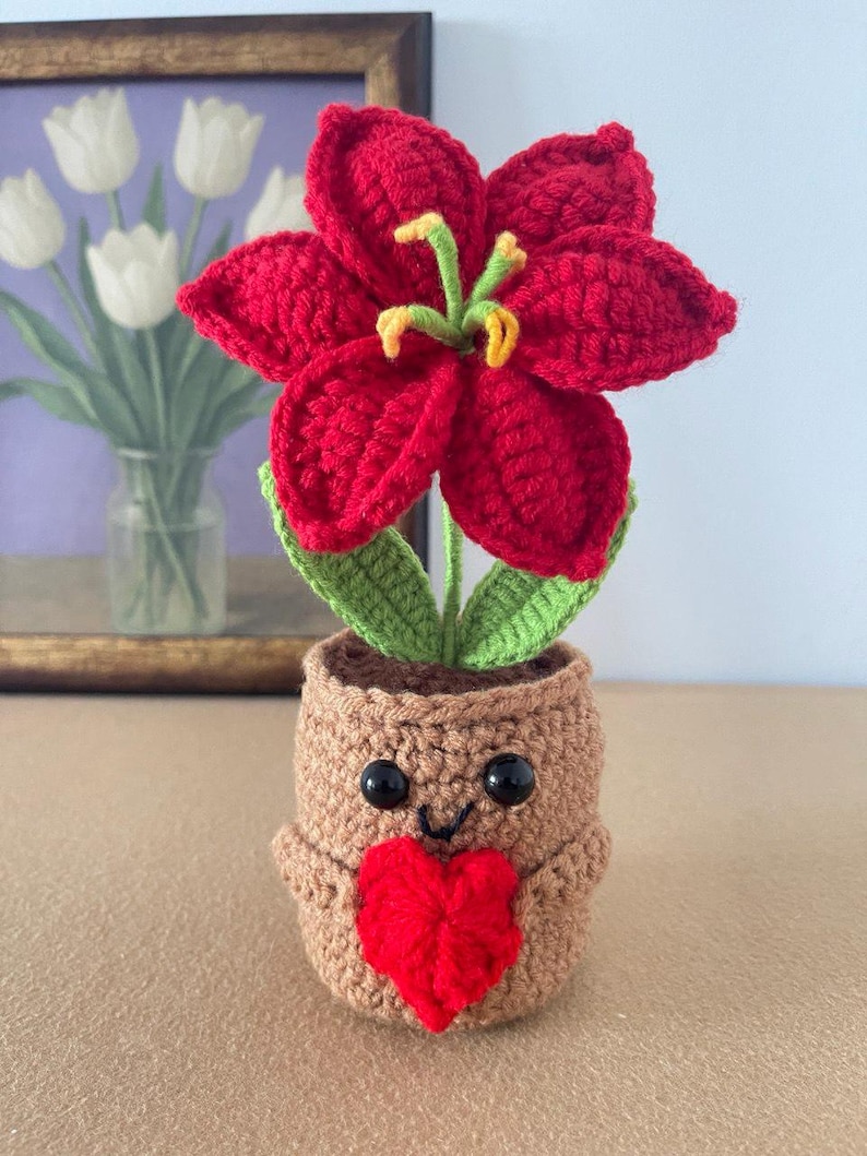 May include: A crocheted red lily flower with green leaves planted in a tan, cylindrical crocheted pot. The pot features two black plastic eyes, a stitched black smile, and a small red crocheted heart attached to the front center.