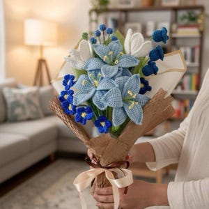 May include: A handcrafted bouquet of crocheted flowers in shades of blue, white, and yellow. The bouquet includes lilies, tulips, and smaller blue flowers, wrapped in brown paper and tied with a cream ribbon. A unique, handmade gift.