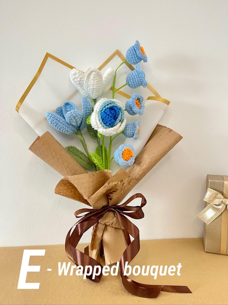 May include: A handcrafted crochet bouquet featuring blue, white, and orange flowers. The bouquet is wrapped in brown paper and tied with a brown ribbon. The text "E - Wrapped bouquet" is visible on the lower left.