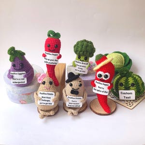Peut inclure: Une collection de peluches de légumes en crochet tenant des pancartes avec des messages positifs. Les peluches comprennent un poivron rouge, une aubergine, une pomme de terre, un brocoli et un chou. Les pancartes ont des messages comme "Tu es un vrai aubergenius !" et "Remplis ta journée de positivité !"