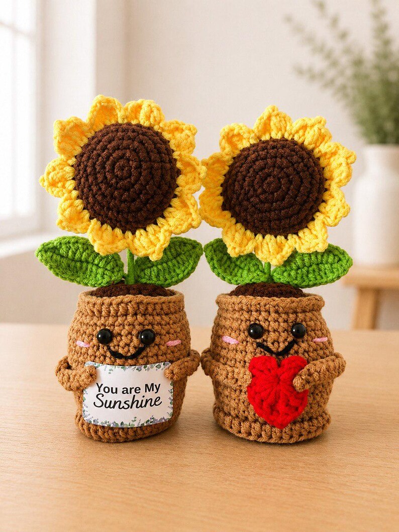 May include: Two crocheted sunflower plants in brown pots with smiling faces and pink cheeks. One pot holds a white sign with the text "You are My Sunshine" and the other holds a red crocheted heart. Each sunflower has yellow petals.