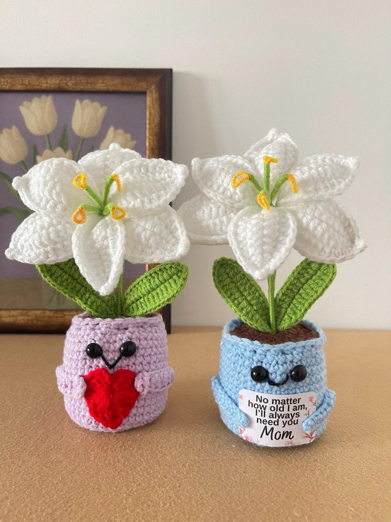 May include: Two crocheted white lily flowers in small pots. One pot is light purple with a red heart, and the other is light blue with a sign that reads: No matter how old I am, I'll always need you Mom. Both pots have black bead eyes and green leaves.