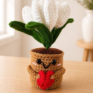 May include: A crocheted amigurumi plant featuring a white flower with green leaves, potted in a tan container with a smiling face, black bead eyes, and pink cheeks, holding a small red crocheted heart against its front with two small arms.
