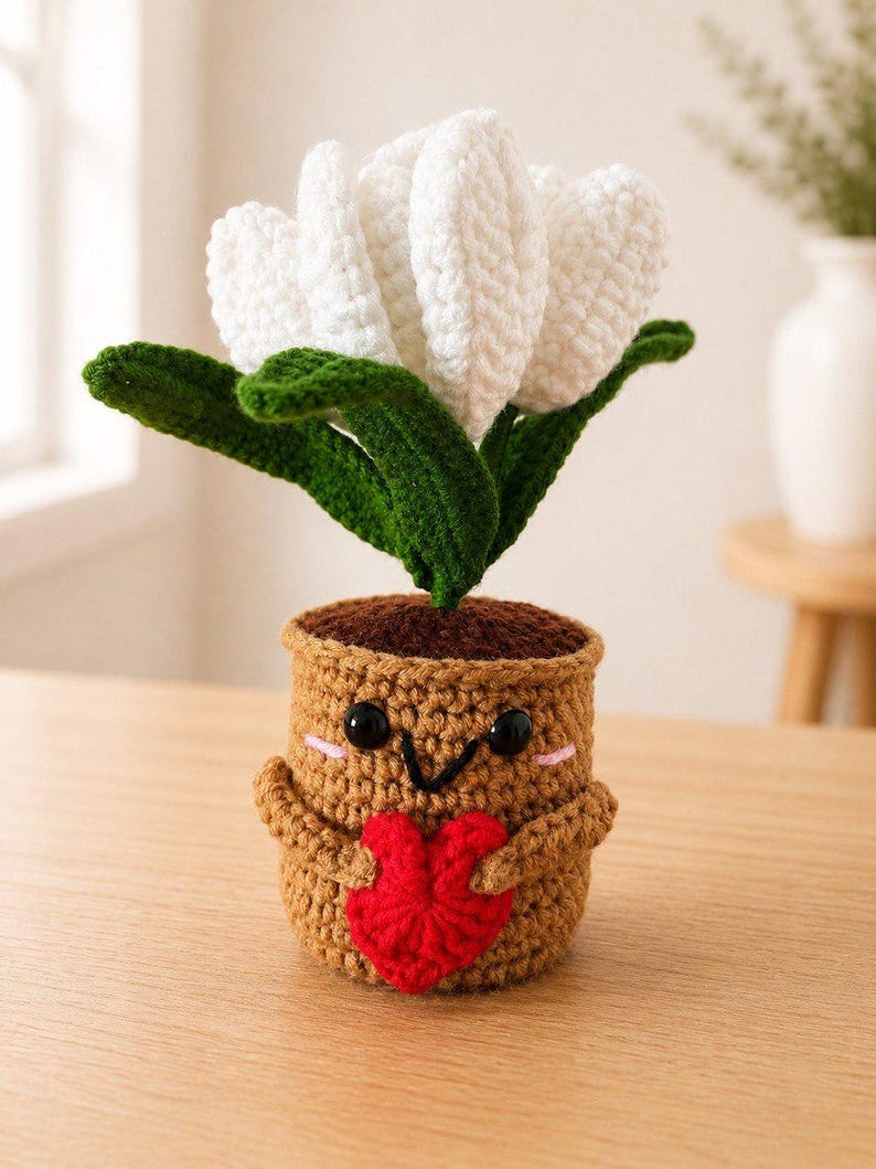 May include: A crocheted amigurumi plant featuring a white flower with green leaves, potted in a tan container with a smiling face, black bead eyes, and pink cheeks, holding a small red crocheted heart against its front with two small arms.