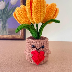 May include: A crocheted orange tulip plant in a light brown pot. The pot features a stitched face with two black bead eyes and a small black smile, and it holds a bright red crocheted heart in its center. The green leaves are visible.
