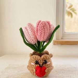May include: A crocheted potted plant featuring three light pink tulip flowers with green stems and leaves. The pot is a round, light brown, textured container with a smiling face, black bead eyes, pink cheeks, and a small red heart held in its center.