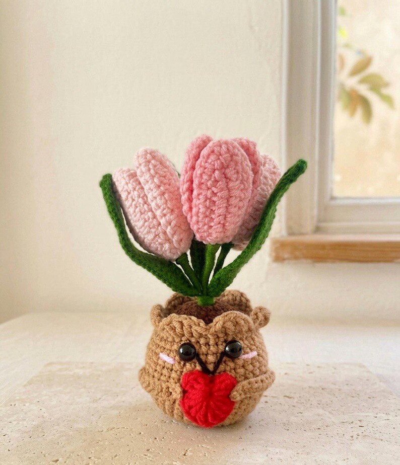 May include: A crocheted potted plant featuring three light pink tulip flowers with green stems and leaves. The pot is a round, light brown, textured container with a smiling face, black bead eyes, pink cheeks, and a small red heart held in its center.