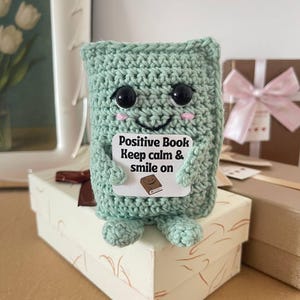 Op de afbeelding: Een lichtgroen gehaakt boekvormig pluche speelgoed met grote zwarte ogen en roze wangen. Het speelgoed houdt een bord vast met de tekst "Positive Book Keep calm & smile on". Het staat op een witte geschenkdoos.