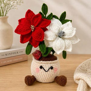 Puede incluir: Una planta en maceta tejida a ganchillo con una flor de pascua roja y otra blanca con hojas verdes. La maceta color crema tiene una cara sonriente bordada con mejillas rosadas y dos pies marrones. Un pequeño regalo rojo está insertado.