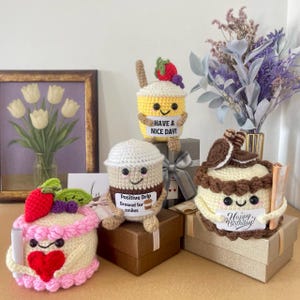 Op de afbeelding: Een verzameling handgemaakte haakartikelen, waaronder een gele beker met aardbeien- en druiventopping, een koffiekopje met een bordje met de tekst "Positive Drip Brewed for smiles" en twee cakes. Eén cake heeft een hart en de andere zegt "Happy Birthday!"