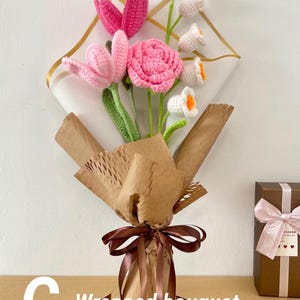 May include: A handmade crochet bouquet featuring pink and white flowers, wrapped in brown paper and tied with a brown ribbon. The bouquet includes pink tulips, a pink rose, and white flowers with orange centers. The text "G - Wrapped bouquet" is visible.