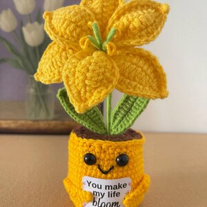 May include: A crocheted yellow flower with green leaves planted in a yellow pot with a smiling face. The pot holds a white rectangular sign with the text You make my life bloom in black lettering. The flower has six petals and a green center.