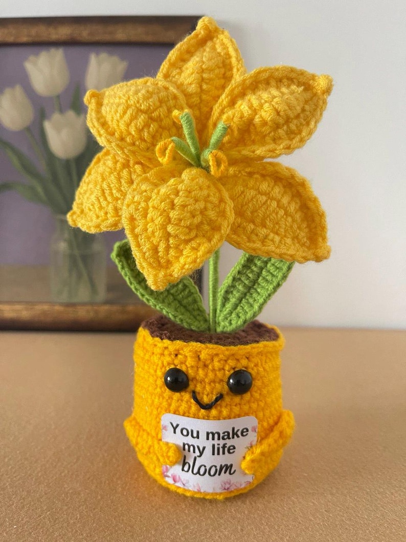 May include: A crocheted yellow flower with green leaves planted in a yellow pot with a smiling face. The pot holds a white rectangular sign with the text You make my life bloom in black lettering. The flower has six petals and a green center.