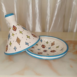 Puede incluir: Una tagine de cerámica blanca con un borde azul y un patrón geométrico colorido. La tapa está quitada y el plato está sobre una superficie blanca.