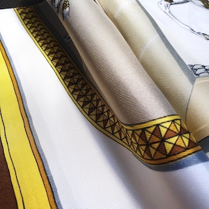 Può includere: Un foulard di seta piegato con base bianca e un bordo geometrico giallo e marrone. Il tessuto presenta una consistenza liscia e lucente con linee di trama diagonali visibili e un'illustrazione dettagliata di un cavallo nell'angolo.