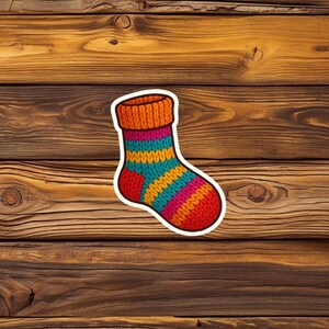 May include: A colourful sticker of a knitted sock with a white border. The sock has an orange ribbed cuff, a striped pattern of pink, teal, and yellow, and a red toe and heel. The sticker is on a wooden background.