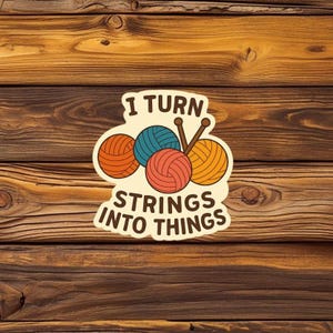 Könnte beinhalten: Ein Aufkleber mit dem Text "I TURN STRINGS INTO THINGS" und Illustrationen von Wollknäueln und Stricknadeln. Die Wollknäuel sind orange, türkis, korallenfarben und gelb. Der Hintergrund ist eine Holzoberfläche.