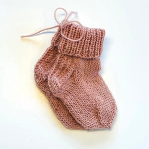 May include: A pair of dusty rose knitted booties featuring a ribbed cuff and a matching yarn tie closure. The booties are crafted with a standard stockinette stitch pattern, showcasing a soft texture and a consistent muted pink colour throughout.