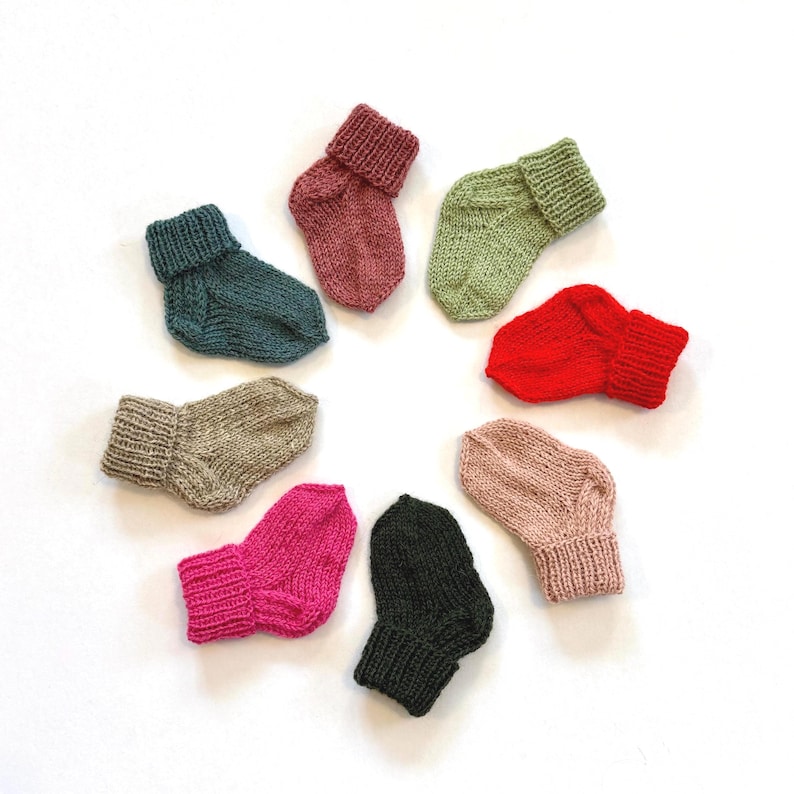May include: A collection of eight small, hand-knitted socks arranged in a circular pattern. The socks feature a ribbed cuff and are available in various solid colours including teal, dusty rose, sage green, bright red, beige, fuchsia, black, and blush.