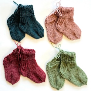 May include: Four pairs of hand-knitted baby socks in teal, dusty pink, deep red, and sage green. Each pair features a ribbed cuff, a stockinette stitch body, and a small yarn tie attached to the top. The socks are arranged in a grid pattern.