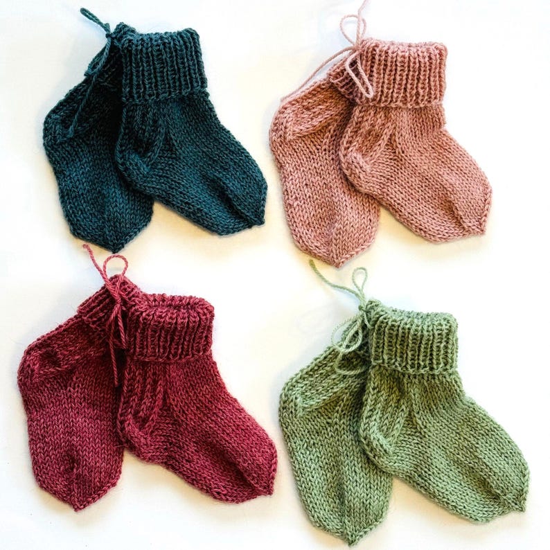 May include: Four pairs of hand-knitted baby socks in teal, dusty pink, deep red, and sage green. Each pair features a ribbed cuff, a stockinette stitch body, and a small yarn tie attached to the top. The socks are arranged in a grid pattern.