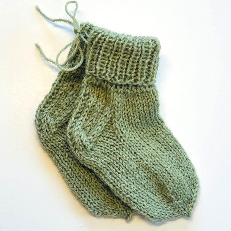 May include: A pair of sage green hand-knitted socks featuring a ribbed cuff and a simple stockinette stitch pattern. The socks are connected by a matching yarn tie at the top, showcasing a soft texture and a classic, functional design.