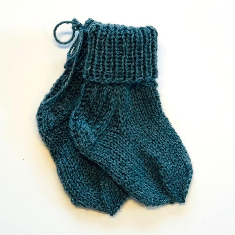May include: A pair of hand-knitted teal socks featuring a ribbed cuff and a classic stockinette stitch pattern. The socks are crafted from a soft yarn and include a small loop of matching thread attached to the top of one cuff for display.
