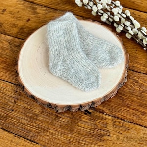 May include: A pair of light grey knitted socks made from a soft, fuzzy yarn. The socks feature a classic ankle-length design with a ribbed cuff and a rounded toe, showcasing a textured, airy knit pattern that appears lightweight and warm.