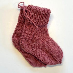 May include: A pair of dusty rose knitted socks featuring a ribbed cuff and a matching yarn tie detail. The socks are crafted from a textured wool-blend yarn with a visible knit pattern, showcasing a folded top edge and a rounded heel construction.