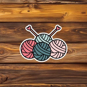 May include: A sticker showing four balls of yarn in pink and teal, with two knitting needles with heart-shaped ends. The design is outlined in black and set against a wooden background, ideal for craft enthusiasts.