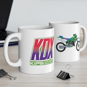 May include: Two white ceramic mugs with a green and white dirt bike graphic on one mug and a purple, red, and green logo that says "KDX KIPS" on the other mug.