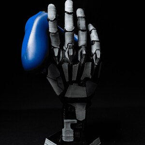 May include: A 3D printed skeletal hand holding a blue video game controller. The hand is posed as if it is holding the controller in a gaming position. The hand is on a hexagonal base.