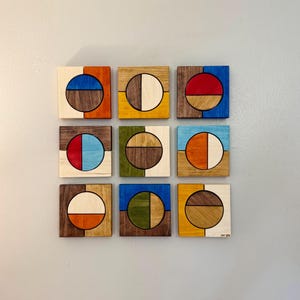 May include: Nine square wooden wall art pieces with geometric designs. Each square features a half-circle and a half-square in various colours, including blue, orange, red, and green, with a natural wood grain finish. The artwork is mounted on a light grey wall.