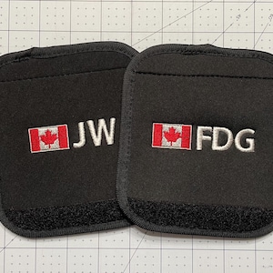 May include: Two black rectangular patches with rounded corners. Each patch features a red and white Canadian flag embroidered, alongside white embroidered initials. One patch reads "JW" and the other "FDG". Each patch has a velcro strip at the bottom.
