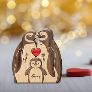 Puede incluir: Figura de familia de ping&uuml;inos de madera con los nombres Jessica, Brian y Janey grabados en ella. Los ping&uuml;inos se besan y la fecha 11.19.22 est&aacute; grabada en un coraz&oacute;n rojo.