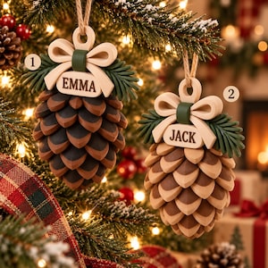 May include: Two pinecone-shaped Christmas ornaments hanging from twine. Each ornament features a cream-coloured bow, dark green pine needles, and a central plaque engraved with the names EMMA and JACK respectively. The pinecones have layered brown and dark brown scales.