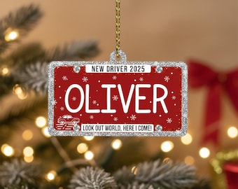 Può includere: Un ornamento natalizio rettangolare con sfondo rosso e bordo glitterato argentato. L'ornamento dice "NEW DRIVER 2025" e "OLIVER" in testo bianco, con fiocchi di neve e una grafica di auto. Il testo in basso recita "LOOK OUT WORLD, HERE I COME!"