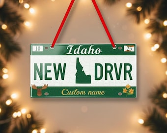 Puede incluir: Un adorno rectangular de matrícula de Idaho con el texto "Idaho", "NEW DRVR" y "Custom name". El adorno es verde con letras blancas y una cuerda roja para colgar. El fondo está borroso con luces navideñas.