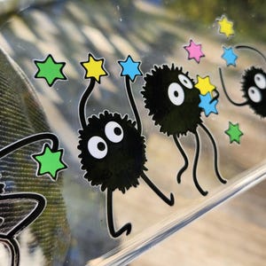 The glass panel showcases playful black cartoon creatures reminiscent of soot sprites, each clutching colorful stars. Stickers of these spirited figures dance across the surface, with a lush forest background visible through the transparent gaps.