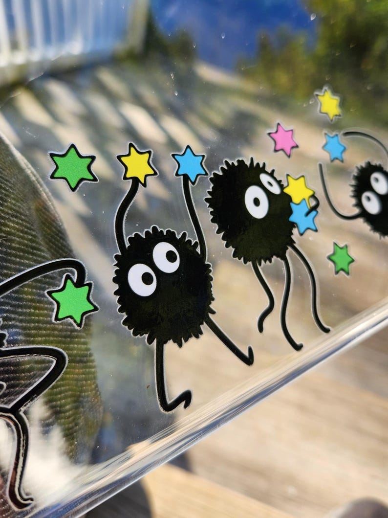 The glass panel showcases playful black cartoon creatures reminiscent of soot sprites, each clutching colorful stars. Stickers of these spirited figures dance across the surface, with a lush forest background visible through the transparent gaps.
