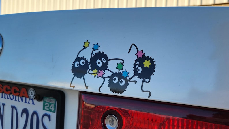 May include: A white car with a black and white sticker of five cartoon characters with stars on their heads. The characters are black with white eyes and are in a playful pose. The car has a Virginia license plate with the number ND205.