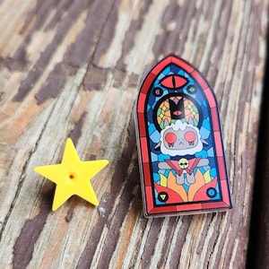 May include: A yellow star-shaped pin and a stained-glass style pin featuring a cartoon character with a red and black border. The stained-glass pin has a red eye, a cat, and a skull design. The background is a wooden surface.