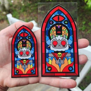 May include: Two stickers featuring a stained-glass design with a cartoon lamb character. The stickers have a red, blue, and yellow color scheme. The lamb has red eyes and is surrounded by flames.