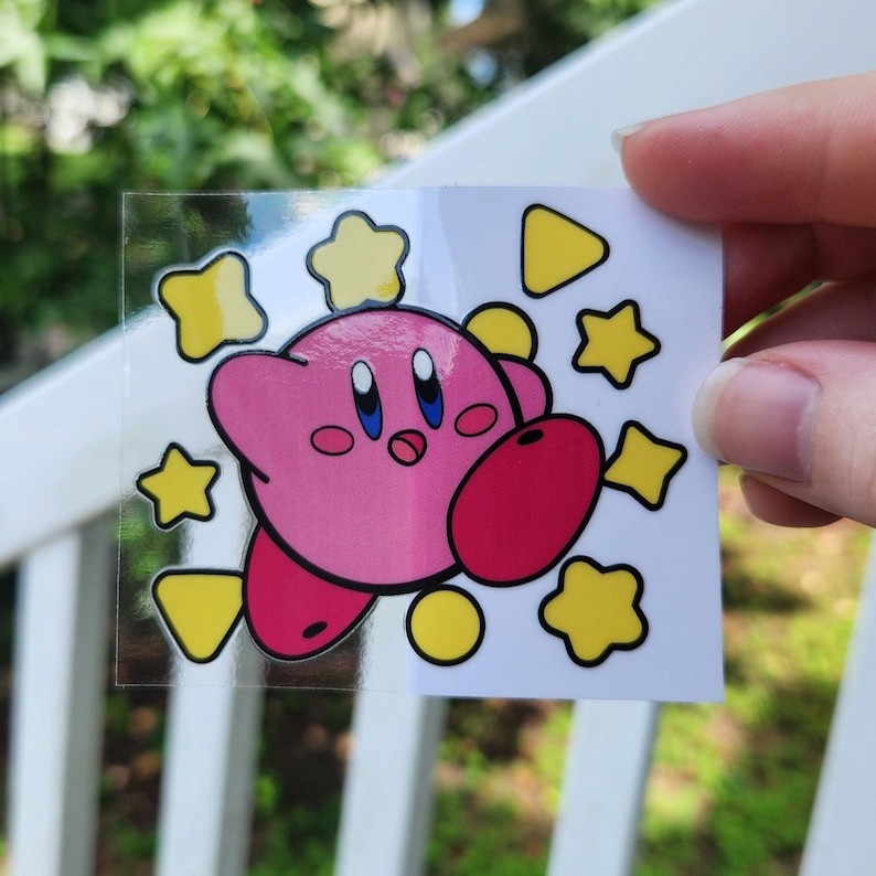 May include: A pink and red cartoon character with big eyes and a surprised expression. The character is surrounded by yellow stars and a yellow triangle. The sticker is on a white background.