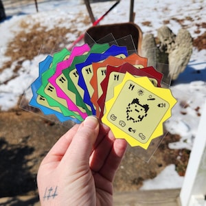 May include: A hand holds a fan of colorful, pixelated game cards. Each card has a unique color, including blue, green, pink, and yellow. The cards feature a pixelated character and various icons. The cards are on clear plastic.