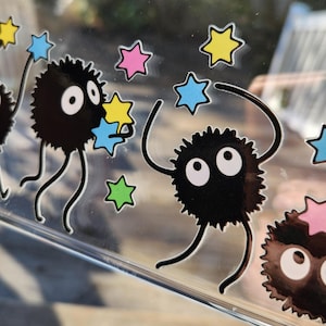 The glass panel showcases playful black cartoon creatures reminiscent of soot sprites, each clutching colorful stars. Stickers of these spirited figures dance across the surface, with a lush forest background visible through the transparent gaps.