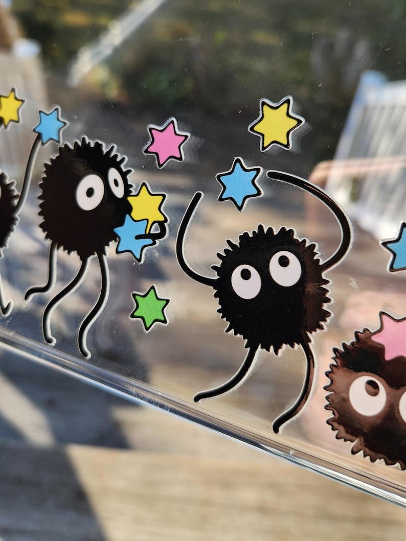 The glass panel showcases playful black cartoon creatures reminiscent of soot sprites, each clutching colorful stars. Stickers of these spirited figures dance across the surface, with a lush forest background visible through the transparent gaps.