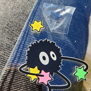 May include: A black and white cartoon character with large eyes and fluffy hair holding a pink, yellow, and green star. The character is surrounded by a black and white outline of a swirling galaxy.