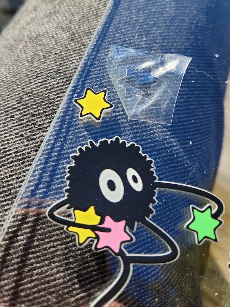 May include: A black and white cartoon character with large eyes and fluffy hair holding a pink, yellow, and green star. The character is surrounded by a black and white outline of a swirling galaxy.