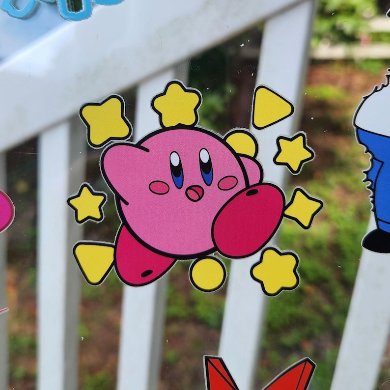 May include: Pink cartoon character with big eyes and a wide smile surrounded by yellow stars. The character is pink with red cheeks and a small red nose. The background is a white surface with a blurry image of a white picket fence.